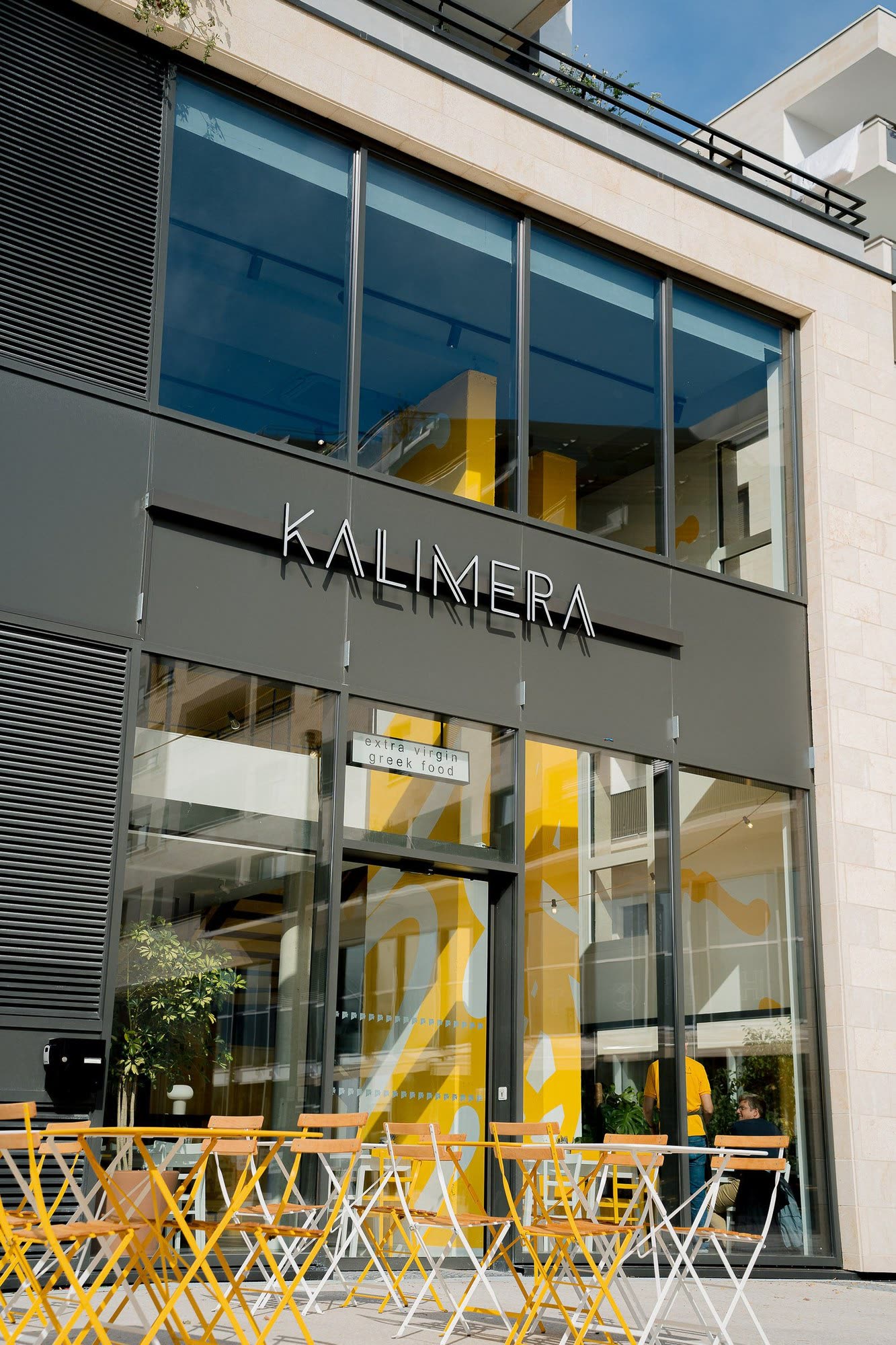 KALIMERA Bordeaux vu de la rue : l’enseigne au-dessus de la façade vitrée et les chaises jaunes de la terrasse