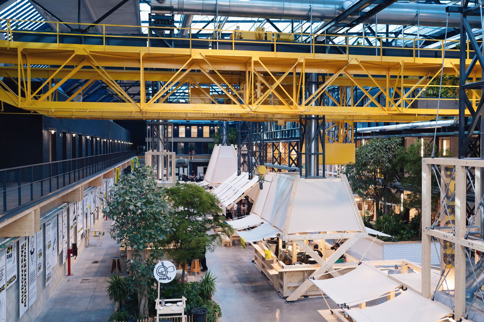 L’intérieur de la Communale, la halle gourmande de Saint-Ouen où KALIMERA a son restaurant : un grand portique d’acier jaune traverse la halle, au-dessus des comptoirs aux toits blancs et des arbres