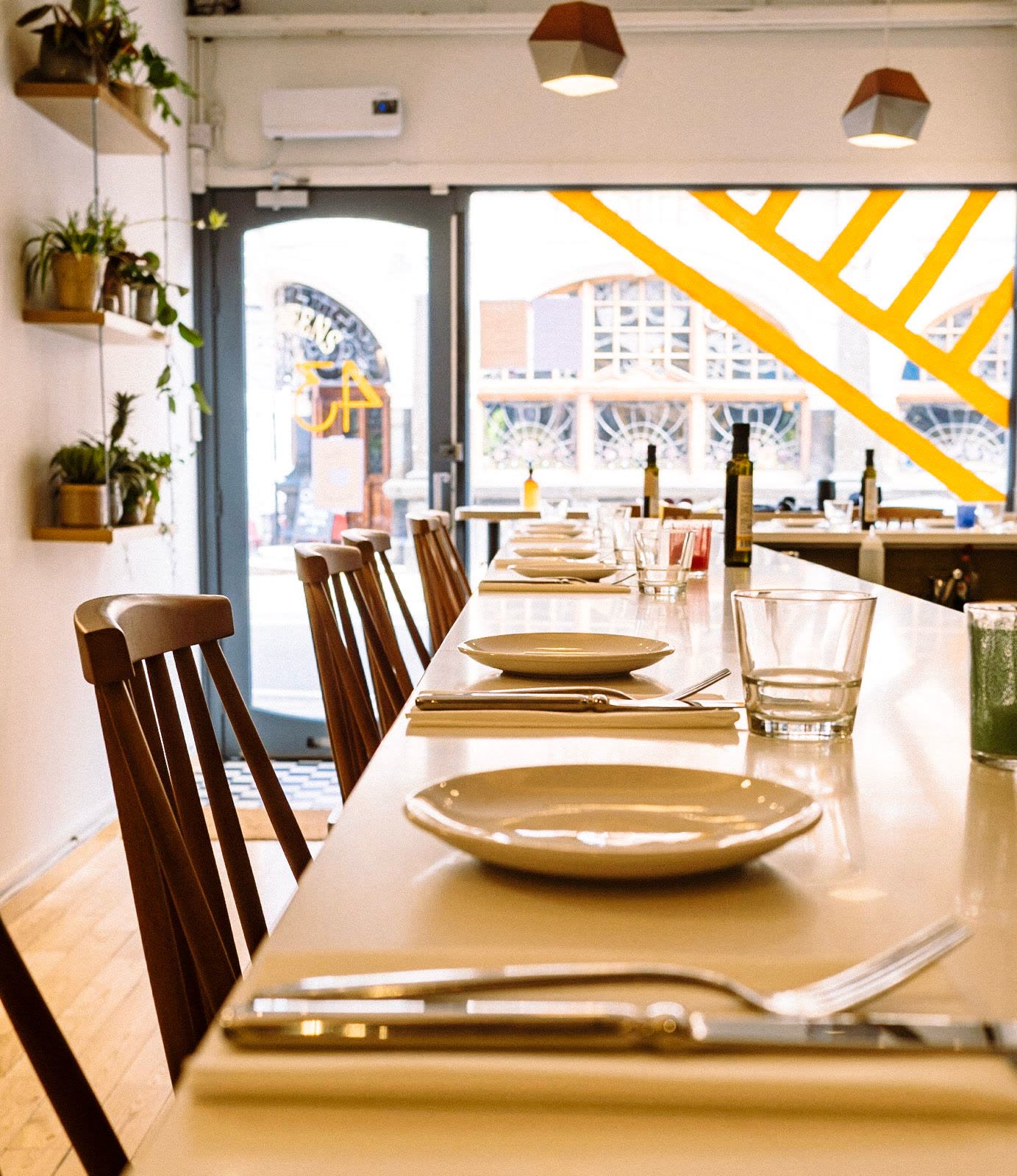 L’intérieur de KALIMERA à Crouch End, à Londres : un long comptoir blanc dressé, assiettes, couverts et verres, des chaises en bois, des plantes au mur et, en travers de la vitrine, les bandes jaunes de la devanture