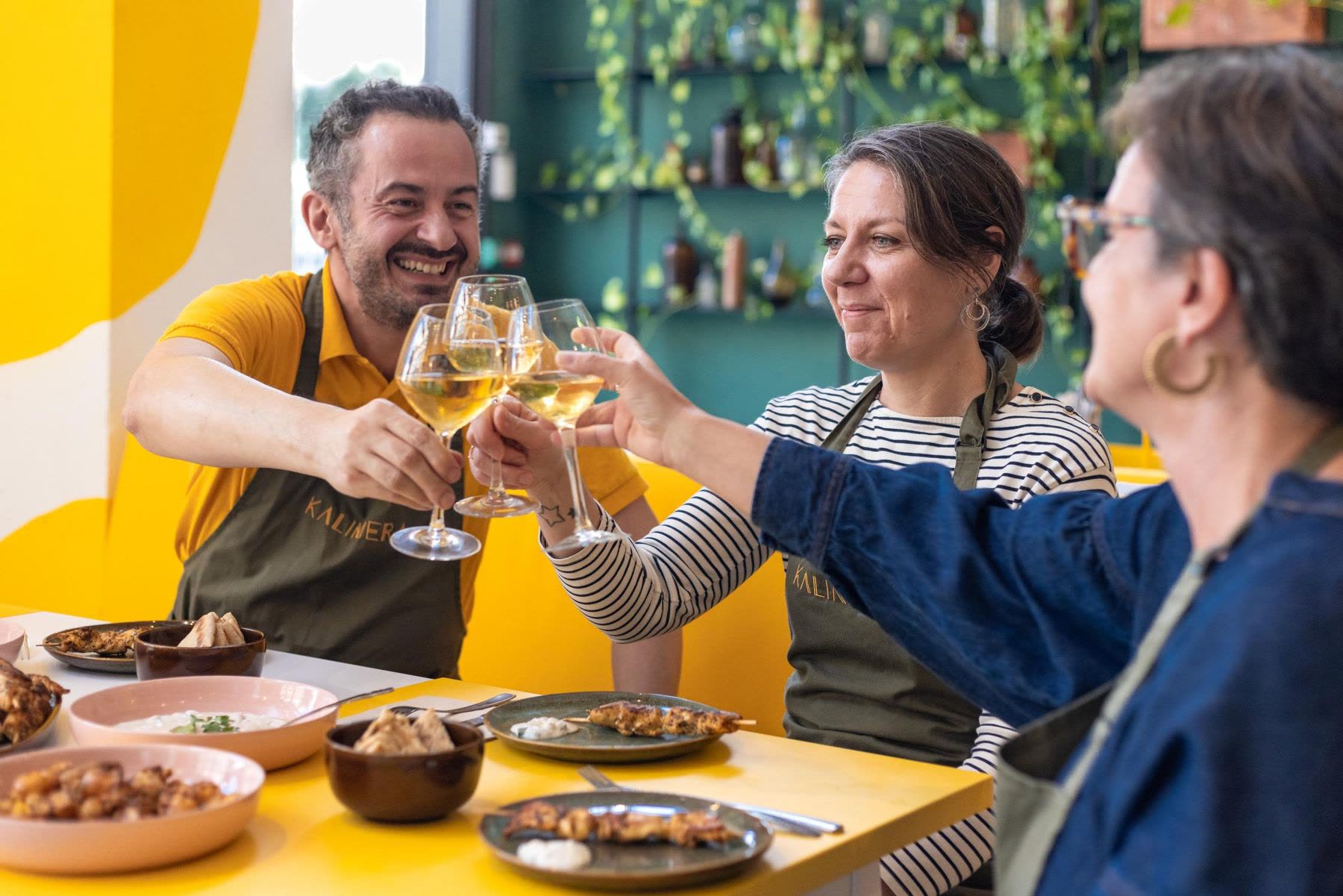 Après un atelier de cuisine KALIMERA à Bordeaux : l’hôte et deux convives trinquent au vin blanc au-dessus d’une table de brochettes de poulet, de tzatziki et de pita
